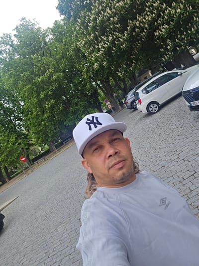 The image is a selfie taken by a man outdoors, likely in Real Sitio de San Ildefonso, Spain, given the contextual information. The man, with light brown or blondish wavy hair visible around his ears and neck, sports light stubble on his face. He is wearing a light grey short-sleeved t-shirt with the "Umbro" logo on the left chest and a white baseball cap with a black "NY" (New York Yankees) emblem on the front. He looks directly at the camera with a neutral expression.
The setting is a paved street or square, featuring grey cobblestones that extend into the background. Along the right side of the frame, several cars are parked. A white compact car is visible behind another white vehicle, whose partial Spanish license plate, "5606 J...", is discernible in the foreground. Further into the scene, a red and white circular traffic sign is visible.
The background is dominated by dense, green foliage from large trees, many of which are adorned with white blossoms, suggesting a spring or early summer season. The bright, natural lighting indicates it is daytime. The overall impression is a casual outdoor moment in a charming, possibly historic, European town.