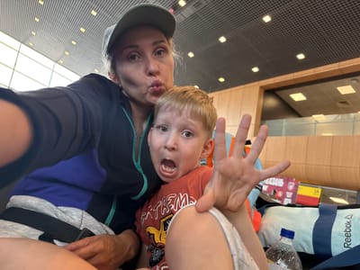 A selfie is taken by a woman and a child. The woman is wearing a black jacket and a green baseball cap. She is making a kissing face. The child, a boy, is wearing an orange t-shirt and is looking at the camera with his mouth open. The background shows a Reebok bag and some boxes. The scene appears to be indoors, possibly at an airport or train station, given the tiled floor and ceiling lights. The overall mood seems playful and lighthearted. The text visible on the bag reads "Reebok".