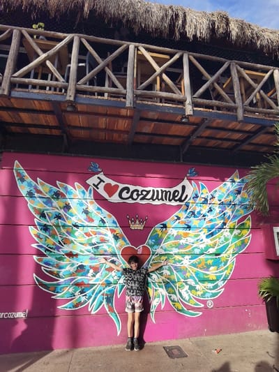 The image shows a young man standing in front of a large pink wall mural in Cozumel, Mexico. The mural depicts a pair of colorful wings with the text "I ♥ Cozumel" and a crown above it. The young man is wearing a black and white shirt and shorts, and is posing with his arms outstretched. Above the mural, there is an open-air structure with a wooden roof and railings. The structure is partially obscured by the edge of the frame. The location, based on the provided coordinates, is approximately near the intersection of Calle 10 and Calle 12 in downtown Cozumel. The scene appears to be captured during the daytime.