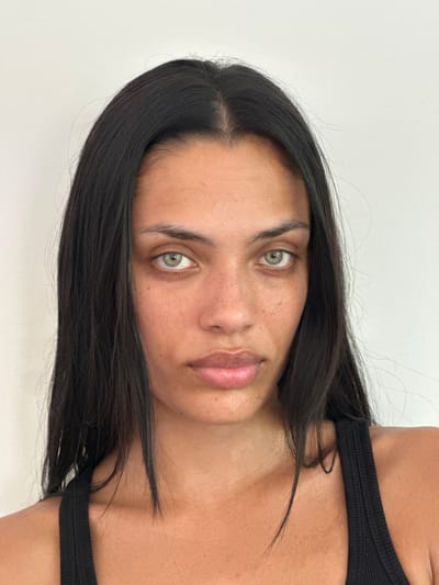 A frontal, medium shot shows a woman with long, dark hair and fair skin with freckles. She has green eyes and is looking directly at the camera. She is wearing a black tank top. The background is a plain, white wall. The image was taken in Le Pré-Saint-Gervais, France.