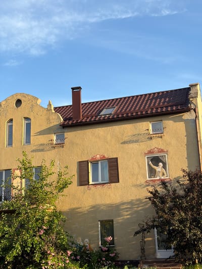 The image shows a yellow, multi-story building with a red-tiled roof, characteristic of architecture found in Slobozhanske, Ukraine. The building has several windows, some adorned with decorative elements. A prominent feature is a painted mural on the facade depicting a person in historical attire waving from a window. Trees and bushes, some with pink flowers, are visible in front of the building, suggesting a garden or yard. The sky is blue with scattered white clouds, indicating daytime. The scene appears serene and captures a piece of the local urban landscape.