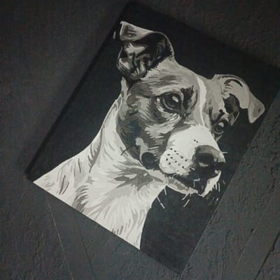 The image displays a painting of a dog's head, rendered in grayscale. The dog is positioned on a dark, textured surface that appears to be a wall. The painting itself is tilted at an angle, adding to the dynamic composition. The dog's face is detailed, with expressive eyes and a discernible snout. The fur is depicted with varying shades of gray, creating a sense of depth and texture. The background of the painting is solid black, which makes the dog's features stand out. The overall impression is that of a striking portrait. The location of this painting in Vyshgorodok, Russia, is not visually evident from the image itself.