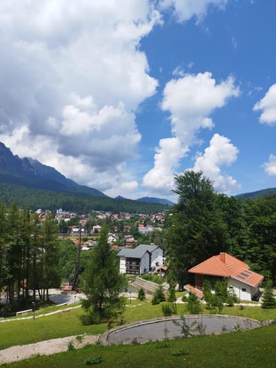 The image captures a panoramic view of the city of Bușteni, Romania, set against a backdrop of majestic mountains. In the foreground, a lush green lawn is dotted with small trees and a paved circular pathway. To the right, a charming white house with a red tiled roof stands prominently, surrounded by greenery.
Further into the scene, a small town unfolds with a cluster of buildings featuring red and grey roofs, nestled amidst dense green trees. In the distance, imposing mountains rise, their peaks partially obscured by clouds. The sky above is a vibrant blue, adorned with fluffy white clouds. A construction crane is visible in the middle ground, suggesting ongoing development in the area. The overall scene conveys a sense of natural beauty and urban life coexisting.