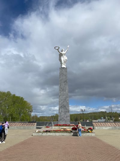 Here's a description of the image:

The image depicts a war memorial in Kirovgrad, Russia. The memorial includes a tall, gray, rectangular monument topped with a white statue of a woman holding a laurel wreath. The monument is set against a backdrop of a cloudy sky. At the base of the monument, there are tiered flower beds filled with red flowers and wreaths. A person is placing flowers on the memorial. On the ground in the lower part of the image, two figures can be seen walking along the brick-paved area. A treeline and buildings are visible in the background, suggesting an urban setting.