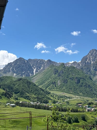 This landscape photograph depicts a mountainous region in Tkarsheti, Georgia. The scene is dominated by imposing, rocky mountains, some of which are partially covered in snow, contrasting with the lush green slopes below. The lower parts of the mountains are densely forested, while higher elevations show bare rock and patches of snow, suggesting a significant altitude.

Nestled on the green slopes is a small village, characterized by a cluster of houses with red and white roofs. A road winds through the village and up the mountainside, hinting at the accessibility of this remote area. A ski lift system is visible in the foreground, with metal poles and cables stretching across the grassy terrain, suggesting the area might be used for winter sports or tourism.

The sky is a clear, vibrant blue, dotted with a few wispy white clouds, indicating fair weather. The sunlight casts shadows on the landscape, suggesting it is daytime, likely late morning or early afternoon. The overall atmosphere is serene and picturesque, showcasing the natural beauty of the Georgian Caucasus. No text is visible in the image.