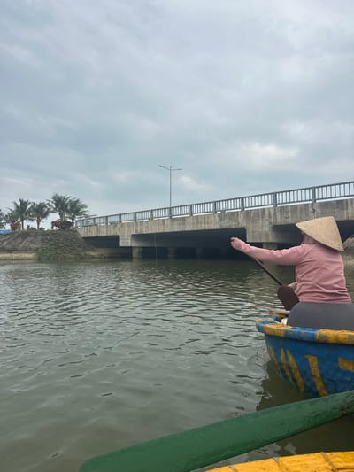 A person wearing a conical hat and a pink long-sleeve shirt leans over the side of a blue and yellow boat, extending their hand towards the water. The boat is positioned near a concrete bridge with railings on either side, visible in the background. The water surface reflects the cloudy sky. Palm trees and vegetation line the shore in the distance, near the bridge support.