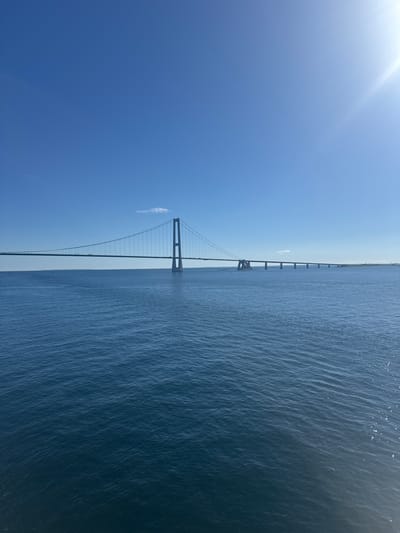 The image depicts a wide shot of a large suspension bridge spanning across a calm, blue body of water under a clear, bright blue sky with some sunlight glinting from the top right corner. The bridge has a prominent central tower and a long deck supported by suspension cables. Several vehicles are visible on the bridge, suggesting it is a functional roadway. The water is a deep blue with subtle ripples, indicating a serene environment. In the distance, land is visible on the horizon. This image captures the vastness of the seascape and the imposing structure of the bridge, likely the Great Belt Bridge in Denmark, connecting the Danish islands of Funen and Zealand. The environment is clear and sunny.