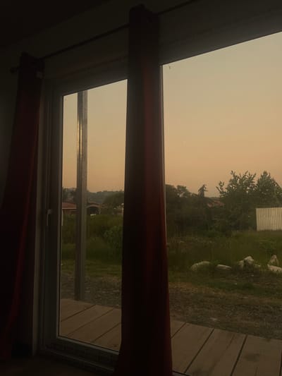 A view through a window shows a hazy, orange-tinted sky, indicative of potential smoke or air pollution. The immediate foreground includes a window frame with a partly visible red curtain. Beyond the window, the landscape reveals green foliage, trees, and a glimpse of buildings in the distance, all obscured by the hazy atmosphere. The presence of trees and distant structures suggests the scene is located in a residential area. The location is Intibucá, Honduras.
