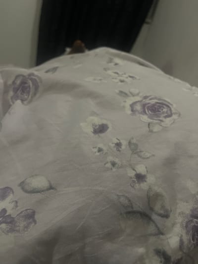 The image shows a close-up view of a light-colored fabric with a floral pattern. The flowers are depicted in shades of purple and gray, with some appearing as smudged outlines. The fabric has a slightly creased texture, suggesting it might be a curtain, upholstery, or bedding. In the background, a dark, textured surface is visible on the left, possibly a wall or a piece of furniture, and a lighter surface below it. The lighting appears to be artificial, creating a soft, diffused illumination across the fabric. No people or specific activities are clearly depicted, and there is no visible text. The overall impression is one of a domestic setting, with the focus on the textile design.