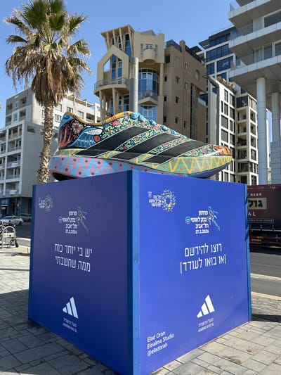 An oversized, intricately decorated Adidas ADIZERO EVO SL running shoe sculpture rests on a solid blue rectangular display base on a paved sidewalk in Tel Aviv-Yafo, Israel. The shoe features a black upper with three white-outlined black stripes, contrasting with vibrant, multi-colored patterns of swirls, floral motifs, geometric shapes, and a wave design across its remaining surface and thick sole.

The blue display base prominently features white and colorful Hebrew and English text and logos. Visible elements include the "TEL AVIV YAFO" logo, "Leumi Bank Tel Aviv Marathon" with the date "27.2.2026", and a stylized runner graphic. Promotional phrases translate to "I have more strength than I thought" and "Run to register (or come to cheer)". The Adidas logo and "The official shoes ADIZERO EVO SL" are displayed, alongside credit to "Elad Oran Einalma Studio @eladoran".

The background reveals a clear blue sky above an urban environment. A tall palm tree stands to the left. A mix of multi-story buildings with varied architectural styles, including a distinctive building with curved elements and large windows, and modern high-rises with glass facades and elevated sections, define the skyline. A street runs parallel to the sidewalk, with a red truck visible in the distance to the right. No individuals are present in the immediate foreground.