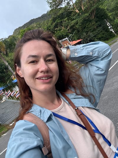 A woman with light brown hair and fair skin is taking a selfie outdoors. She is smiling and has her right arm raised behind her head. She is wearing a light blue button-up shirt over a beige t-shirt, and a brown bag strap is visible across her chest. In the background, there is a road, lush green trees on a hillside, and several flags. The flags appear to be Thai flags, suggesting the location is in Thailand. The environment suggests a tropical or sub-tropical climate. There is a slight overcast sky. The scene is captured from a slightly low angle.
