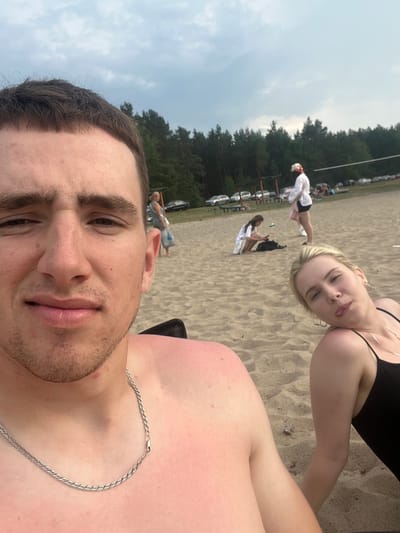 A selfie is taken on a sandy beach with trees and a cloudy sky in the background. The photo is close-up, with the face of a young man in the foreground, looking directly at the camera. He has short brown hair and is wearing a silver chain. To his right, and slightly further back, is a young woman lying down and looking away from the camera with her tongue sticking out. She is wearing a black swimsuit. In the background, other people are visible walking and sitting on the beach, and cars are parked near the treeline. There appears to be a volleyball net set up in the distance. The location is stated to be Загародскі сельскі Савет, Belarus.