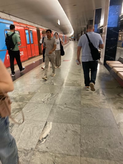 In a subway station setting, several individuals are present. The main subject is a person in the foreground, seen from the back, wearing a light blue t-shirt and dark pants, with a black bag slung across their chest. They appear to be holding a white object, possibly a bottle.
Further down the platform, a group of people are visible. One person in a grey t-shirt is looking towards the camera, while another in a white t-shirt is standing next to them. Behind them, other individuals are partially obscured. A red subway train is stopped at the platform, its doors open. The station's floor is tiled with a granite-like pattern.
The image is taken from a tilted perspective, making it appear as if the people and train are standing on a vertical plane. The lighting suggests an indoor environment, typical of a subway. There are no explicit clues about the time of day or weather, but the setting itself implies an urban underground transit area.
Visible text on the train, partially obscured by the image's orientation, appears to be in Cyrillic script, including the letters "СЯ!". The overall mood seems casual, with people waiting for or disembarking from the train.