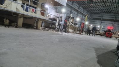 Inside a large, industrial building, a massive, light-colored machine dominates the left side of the frame. Multiple workers are positioned around the machine, appearing to be engaged in operating or maintaining it. A red forklift is visible on the right, with what seems to be a pile of wood behind it. The lighting is a combination of natural and artificial, casting shadows across the concrete floor.