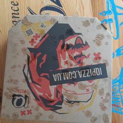 The image shows a close-up, overhead view of a pizza box. The box is made of cardboard and has a design printed on it featuring a stylized, angular illustration in red, black, and yellow, resembling a cartoonish face or character. There are brown stains, possibly from grease or food, scattered across the surface of the box. In the upper left corner, a QR code is visible. Below the QR code, there are two red 'x' marks. Text that reads "10PIZZA.COM.UA" is printed on a black rectangle across the middle of the box. To the left of the main illustration, the word "PIZZA" is printed vertically, and below it is a logo that appears to be a stylized 'a' or 'o'. The pizza box is placed on a wooden surface with visible grain. In the background, there are elements of blue graffiti or painted designs on the wooden surface. The image does not provide any information or visual cues to suggest a location in Dubrivka, Ukraine.