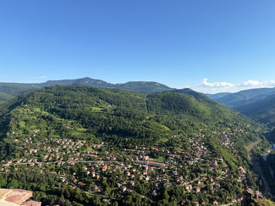 This is a wide-angle, aerial view of the village of Milanovo, Bulgaria, situated within a lush, mountainous landscape. The scene is set on a clear, sunny day with a vibrant blue sky dominating the upper portion of the frame. Rolling green hills, densely covered with trees, form the backdrop and surround the village, which is nestled in a valley.
The village itself is characterized by a cluster of houses with red-tiled roofs, interspersed with trees and winding roads. The architecture appears traditional, with many of the buildings being relatively small and residential. A notable structure, possibly a school or community building, with a larger, more rectangular design and a sports field, stands out in the center of the village. The topography is clearly steep, with houses built on the slopes, creating a picturesque and somewhat precarious setting.
To the right of the village, a river flows through the valley, and parallel to it, a railway line can be seen, indicated by the tracks and a service road. In the foreground, the edge of a stone structure, possibly a wall or a viewpoint, provides a tactile element and suggests the vantage point from which the photograph was taken. The overall impression is one of a peaceful, rural community embraced by the grandeur of nature. The time of day appears to be late morning or early afternoon, given the bright sunlight and the clear visibility. No people are clearly visible, and no text is present in the image.