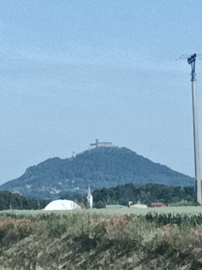 The image shows a wide shot of a rural landscape with a hill topped by a structure in the background. In the foreground, there is a field of vegetation, with some red flowers visible. In the middle ground, there are some buildings, including what appears to be a white dome and a tall, slender spire. A power pole is prominently visible on the right side of the frame. The sky is a clear, light blue. The scene is likely located in Bělá pod Bezdězem, Czechia, given the context of the surrounding hills and architectural styles.