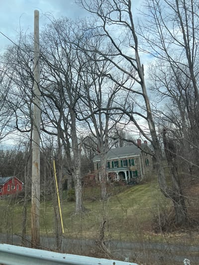 This image depicts a static outdoor scene in the Town of New Windsor, United States of America, characterized by a transitional season, likely late winter or early spring.
In the immediate foreground, a metallic, light gray guardrail is partially visible, suggesting a roadside perspective. Beyond the guardrail, a dark, paved surface, possibly a road or driveway, is discernible.
The midground is dominated by numerous bare deciduous trees, their skeletal branches extending across the frame and obscuring parts of the landscape. A tall, light-colored wooden utility pole stands vertically on the far left, rising high towards the sky. A bright yellow vertical utility marker or cable is present to the right of the utility pole, partially hidden by tree branches.
To the left, set back among the trees, a red building with multiple rectangular windows is partially visible, consistent with a barn or a smaller auxiliary structure. To the right and elevated on a grassy slope, a two-story historical stone house is the central architectural feature. This house features a gray shingled roof, two visible red brick chimneys, and multiple windows fitted with dark green shutters. A white, multi-columned porch extends across the front facade of the house. A dark metal fence delineates a portion of the property in front of the stone house. The ground is covered with patchy, mostly dormant green and brown grass, with sparse undergrowth.
The background consists of more bare trees receding into the distance, under an overcast sky filled with gray and white clouds. No individuals are present within the scene.
