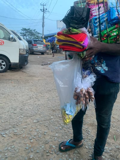 An individual stands in a gravelly urban environment, likely a marketplace in Elelenwo, Nigeria, carrying a large assortment of goods. The person is wearing dark pants and blue flip-flops. They are holding a large bundle of colorful cloths in plastic wrap, along with packages of snacks and face masks. To the left of the individual, two white vans are parked, and a silver SUV is visible in the background. The sky is overcast, suggesting either early morning or late evening. The scene depicts a street vendor engaged in their trade amidst everyday urban activity.