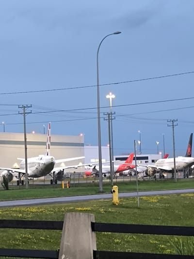 The image depicts an outdoor scene at an airport, likely the Montréal–Mirabel International Airport in Mirabel, Canada, under a twilight sky. Several commercial airplanes are parked in the foreground and middle ground. On the left, a large white airplane with a distinctive red and white striped tail is prominently displayed. To its right, a red and white airplane is visible, and further to the right, another white airplane with a dark tail featuring a red maple leaf logo is parked. In the background, a large building, possibly a hangar or terminal, is situated behind the aircraft. The environment is characterized by a grassy area with scattered yellow flowers, a paved road, and utility poles with overhead wires. A bright yellow fire hydrant stands near the road. The lighting suggests it is either dusk or early morning. There are no visible people in the frame.