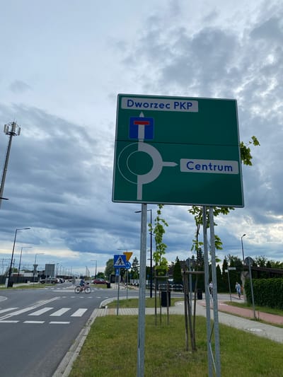 This daytime image, set in Dobieżyn, Poland, captures a street scene under an overcast sky with glimpses of blue. The primary subject is a large green directional sign positioned on the left side of a road, which leads to a visible roundabout.

The green sign displays white text, oriented vertically. On its left side, it reads "Dworzec PKP", and on its right side, "Centrum". A white roundabout symbol with an arrow pointing towards "Centrum" indicates it as an exit from the roundabout. To the left of the roundabout symbol, a blue and red "no entry" traffic sign with a left-pointing arrow suggests a specific turn or route for "Dworzec PKP".

On the right side of the road, a person is seen cycling on a paved surface, appearing to be near a designated pedestrian crossing marked by white stripes. Further along the road, a blue square sign with a white triangle depicting a walking person (indicating a pedestrian crossing) is visible, accompanied by a yellow triangular warning sign above it.

The scene includes elements of urban infrastructure: streetlights, metal fences, and several newly planted trees with support stakes lining the road. Grassy areas and paved pedestrian paths flank the roadway. In the background, residential buildings with hedges and more trees are discernible. A tall cellular tower stands against the cloudy sky in the far left. The overall impression is a functional, organized streetscape in a suburban setting.