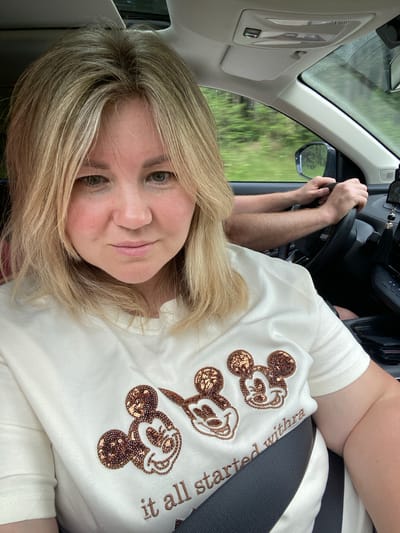 A selfie taken from inside a car shows a woman with blonde hair and green eyes looking at the camera. She is wearing a white t-shirt adorned with three Mickey Mouse heads made of sequins and embroidered text. The text on the shirt reads "it all started withra". A man's arms are visible, one of which is steering the car. The background shows trees and greenery, suggesting the car is in a rural or wooded area. The interior of the car is visible, including the dashboard and a sunroof. The image does not provide any information about the city of Abdes-Urdes, Russia.