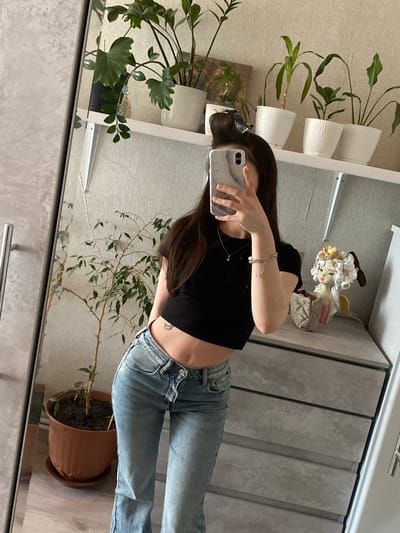 A young woman with dark hair, pulled back into a bun, stands in front of a mirror and takes a selfie with her phone. She is wearing a black crop top and light-wash, high-waisted jeans. She has a small crescent moon tattoo on her hip. In the reflection of the mirror, there are several potted plants on a shelf above a gray dresser. A decorative figurine of an angel is placed on the dresser. The room appears to be a bedroom or dressing area, with a gray wardrobe to the left of the mirror. The floor is wooden. The scene does not provide any specific visual clues that would allow for a definitive location in Nizhnyaya Maktama, Russia.