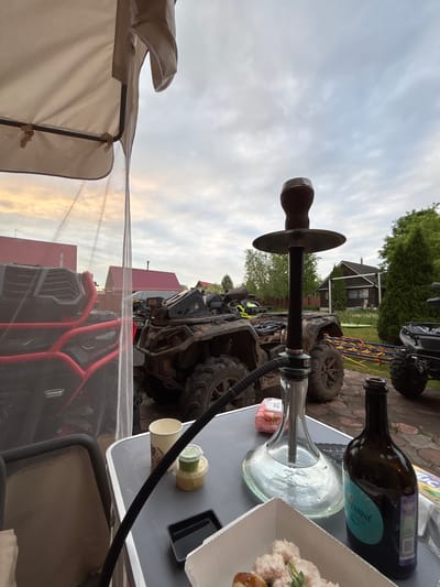 A hookah sits on a table outdoors in Ogryzkovo, Russia. The table also holds a bottle of wine, a container of food, and a small jar. Multiple all-terrain vehicles are visible in the background. A tan canopy is partially visible in the lower left. The sky is overcast.