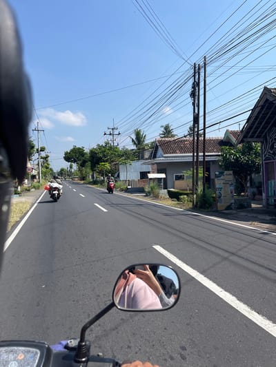 The image depicts a view from the perspective of someone riding a motorcycle on a paved road in Purworejo, Indonesia. The road is lined with buildings and vegetation, with power lines overhead. In the foreground, a motorcycle's handlebar and side mirror are visible. The side mirror reflects the rider wearing a pink hoodie and the back of a person in a grey shirt. Two other motorcycles are seen ahead on the road; one is carrying a red and white package, and another is red. A sign for "HUANG COFFE MAKANAN & MINUMAN" is displayed on the right side of the road. The sky is clear and blue.