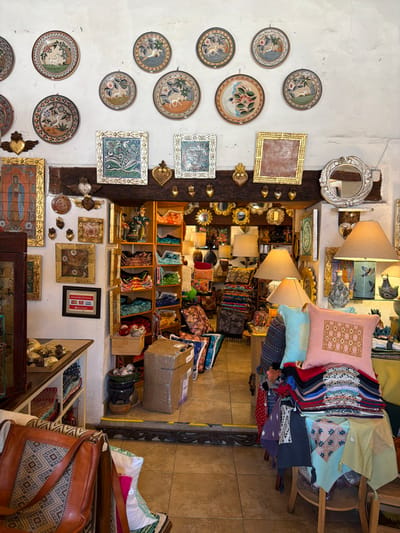 This image captures the interior of a richly decorated artisanal shop, filled with various handcrafted goods and home decor items. The perspective is slightly angled downwards, looking into a densely packed retail space. The image is vertically oriented (if viewed in its original state) but depicts a horizontal scene if rotated clockwise by 90 degrees, revealing a wall on the left, display shelves and tables in the center, and a tiled floor on the right.
On the left side, a light-colored, possibly white or off-white, plastered wall serves as a backdrop for numerous decorative ceramic plates. These plates, predominantly round, are arranged in an informal grid pattern and feature intricate hand-painted designs, often depicting classical or mythological figures (like women's profiles), botanical motifs, and floral patterns in earthy tones with touches of blue, green, and orange. Some square and rectangular framed items, one a reddish-brown with a golden frame and others with blue and white intricate tile-like designs, are also hung on this wall. Near the top, a silver-colored, ornate mirror with a carved frame reflects a portion of the shop's interior.
A prominent, thick dark wooden beam runs vertically down the wall, separating the plastered section from the main display area. This beam is adorned with several small, golden, heart-shaped decorative pieces, along with a larger, more elaborate heart-shaped carving.
The central part of the shop is a vibrant display of merchandise. Two lamps with light-colored, bell-shaped fabric shades emit a warm glow, illuminating the diverse collection of goods. Shelves and tables are heavily laden with items. A stack of colorful cushions and pillows is visible, including a prominent pink pillow with a delicate golden geometric pattern and a light blue one underneath. Other pillows feature stripes, floral designs, and various patterns.
Below the lamps and pillows, several wooden shelves hold an array of small sculptures or figurines. These appear to be elongated, stylized forms, possibly human or animal, rendered in various vibrant colors like teal, green, orange, and blue. Interspersed among these are numerous small, ornate, gold-framed mirrors of different shapes (round, oval, rectangular), adding a touch of antique charm. Other framed artworks, including one at the lower center depicting what appears to be a religious figure in blue and gold, are also displayed. A small green figurine, possibly a frog or monster, can be spotted on one of the shelves.
Towards the mid-right, a brown cardboard box, sealed with tape, sits on a shelf or surface, somewhat out of place amidst the artistic goods. Below it, more shelves are filled with neatly folded textiles, fabrics, and possibly clothing items in a variety of patterns and colors.
The right side of the image reveals a light brown or beige tiled floor. A wooden chair or small table, partly visible in the far right, is draped with colorful textiles and more pillows. In the bottom right corner, a large leather bag with an embroidered or woven patterned panel suggests that accessories are also sold here. A dark, intricately carved wooden element, possibly part of a doorway or large furniture, stands against the tiled floor.
The overall impression is that of a cozy, slightly cluttered, but well-curated shop specializing in handcrafted goods, home decor, and possibly fashion accessories, with a rich, inviting atmosphere created by the warm lighting and abundance of unique items. No discernible text is clearly visible in the image.