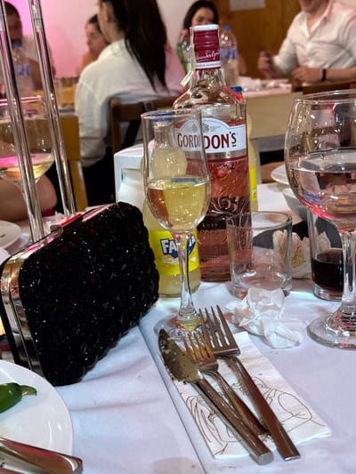 The image depicts an indoor dining or social gathering in Debrashtitsa, Bulgaria. The foreground features a white tablecloth with various items arranged upon it. A black, jewel-embellished clutch bag with silver-toned hardware is centrally positioned on the left. To its right, three pieces of silverware (two forks and one knife) rest on a white paper napkin adorned with a faint graphic of figures.
Multiple glasses and bottles are present on the table. A stemmed glass containing a yellowish liquid is visible on the left, partially obscured by two clear, vertical tubular elements, likely part of a table centerpiece. Next to this, a partial yellow bottle labeled "FANTA" is visible. A prominent bottle of "GORDON'S PREMIUM PINK DISTILLED GIN" with a red cap and pink liquid is centrally located. To the right of the gin bottle, an empty, clear, short tumbler glass sits beside a crumpled white napkin. Further right, a stemmed glass holds a dark liquid, and another stemmed glass contains clear liquid (water). A small section of a white plate with a green chili pepper is visible in the bottom-left corner.
The background is out of focus, revealing several individuals seated at tables. In the mid-left background, a person with dark hair, wearing a white collared shirt, is seated with their back partially to the camera. In the mid-right background, a male individual wearing a light-colored collared shirt is visible, with his hands positioned on the table. Other blurred figures and table settings are discernible throughout the background. The lighting in the background exhibits a pinkish hue, possibly from ambient illumination. A wooden chair back is visible in the mid-ground.