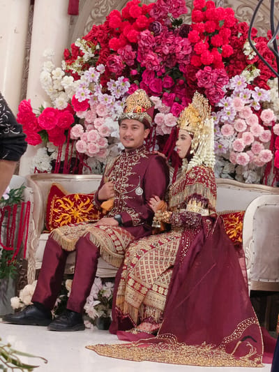 In Meuliek, Indonesia, a newlywed couple, adorned in traditional maroon and gold attire, is seated on a white couch. The groom wears a bejeweled crown and a richly embroidered tunic and pants, while the bride sports an elaborate golden headdress and a matching embellished gown. They are surrounded by a vibrant backdrop of artificial flowers, predominantly in shades of red, pink, and white. The groom is seen looking to his right, with his left hand resting on his lap. The bride, with her head tilted to the left, appears to be looking away from the camera. The scene is likely a wedding ceremony or reception, set within an indoor venue, suggested by the presence of ornate pillars and wall decorations.