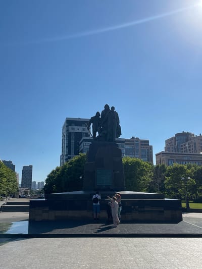 The image depicts a monument with a group of bronze figures on a dark stone pedestal. Several people are visible near the monument, some standing and looking at it, while others are seated or walking nearby. The setting is urban, with modern buildings and lush green trees visible in the background. The sky is clear and blue, with a bright sun causing lens flare in the upper left corner, suggesting it is daytime. The overall atmosphere appears relaxed and casual, with people enjoying the outdoor space. There is no visible text in the image.