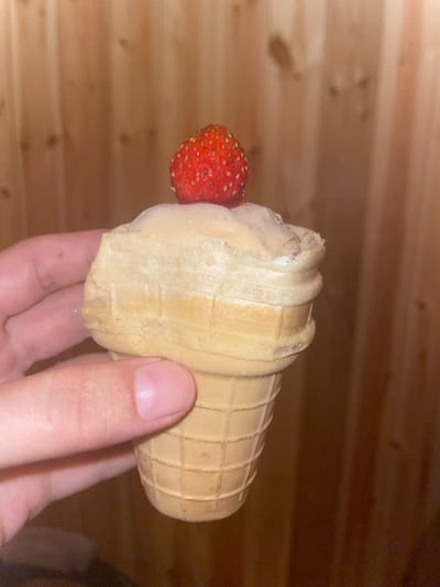 A close-up, eye-level shot shows a person's hand holding a small, light-colored ice cream cone topped with a single strawberry. The ice cream is a pale beige color and has a slightly rough texture, with some glistening from condensation or sugar. The waffle cone is light brown and has a ridged pattern. The strawberry is bright red and has visible seeds. The background is out of focus, showing vertical wooden planks. The image appears to have been taken indoors, likely in a home setting, with the wooden background suggesting a wall or furniture. There is no information in the image to suggest a specific location in the city of Маминское, Russia.