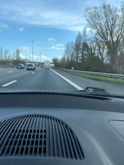 The image depicts a forward-facing view from inside a vehicle traveling on a multi-lane highway near Temse, Belgium. The foreground prominently features the vehicle's dark grey dashboard, textured with a fine pattern. A circular speaker or ventilation grille with vertical and horizontal slats is visible on the lower left of the dashboard. Further up, near the base of the windshield, is a narrow horizontal air vent. A black windshield wiper arm and blade assembly are visible on the lower right of the windshield.
Ahead, the highway comprises at least three lanes of asphalt pavement marked by white dashed lines, indicating traffic moving in the same direction. A solid white line defines the right edge of the road, bordering a paved shoulder. Several vehicles, predominantly passenger cars and a distant white box truck, are traveling on the highway.
The immediate right side of the road features a metal guardrail, beyond which is a strip of grass followed by dense vegetation, consisting mainly of bare deciduous trees and some evergreens, suggesting a winter or early spring season. Similar bare trees line the left side of the highway in the background. Utility poles or light standards are visible on the left, and a bridge or overpass structure spans the highway in the far distance. The sky above is partly cloudy with significant areas of clear blue.