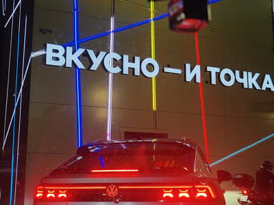 In Gorki-2, Russia, at night, a building facade is illuminated by vertical blue and white lights, with horizontal blue and yellow lights crossing the signage. The building displays white, three-dimensional lettering spelling "ВКУСНО – И ТОЧКА" in Russian, which translates to "Tasty - and that's it." In the foreground, the rear of a dark-colored SUV is visible, its brake lights illuminated red. To the right, a person on a motorcycle wearing a helmet is partially visible, with a license plate reading "4243 BP/77". The overall scene appears to be a commercial establishment, possibly a fast-food drive-through, given the presence of vehicles and prominent signage.
