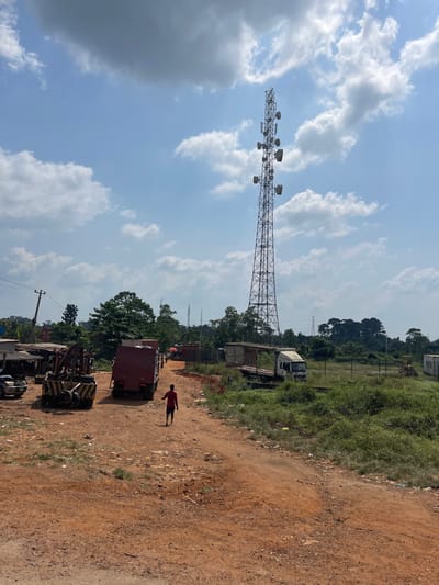 The scene in Oko-Eko, Nigeria, depicts an unpaved, reddish-brown dirt road under a clear sky with scattered white cumulus clouds. A tall metal lattice telecommunications tower, equipped with multiple dish antennas and ancillary equipment, dominates the right half of the frame, extending into the sky.

In the foreground, a single individual, appearing male, dressed in a red short-sleeved shirt and dark shorts, walks away from the camera on the dirt road, roughly center-left. To the immediate left of this individual, a large red box truck is parked, facing away from the camera. Further left, a yellow and black heavy-duty vehicle, consistent with a tow truck or mobile crane, is stationary. Beyond these vehicles, several low-rise structures, possibly stalls or workshops, are visible, along with a utility pole supporting overhead lines.

To the right of the walking individual and the dirt road, an open, unkempt grassy area with sparse vegetation contains a white flatbed truck parked on a small elevated platform. Further in the background, another piece of yellow heavy machinery, possibly an excavator or bulldozer, is partially visible. A line of green trees forms a distant horizon, and several large metal electricity pylons are discernible against the background sky. The overall environment appears semi-rural or industrial.