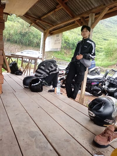 A young man in a black jacket with white stripes stands in an open-sided wooden shelter with a concrete floor, in the vicinity of Jalan Raya Pangalengan - Bandung, Kecamatan Pangalengan, Indonesia. The man is looking to the left. He wears black pants and a dark satchel. He is standing in front of several motorcycles parked just outside the shelter, two of which are partially visible. On the floor next to him are two black helmets and a black backpack. A person is sitting on the ground, partially obscured, wearing a striped black and white sweater. A partially visible silver car and lush green vegetation can be seen outside the shelter. A sign in Indonesian hangs on the wall of the shelter. A small, clear plastic water bottle lies on the floor in front of the motorcycles. The floor of the shelter is made of wooden planks. The ceiling is open to the sky and is constructed of wooden beams. The weather is cloudy.