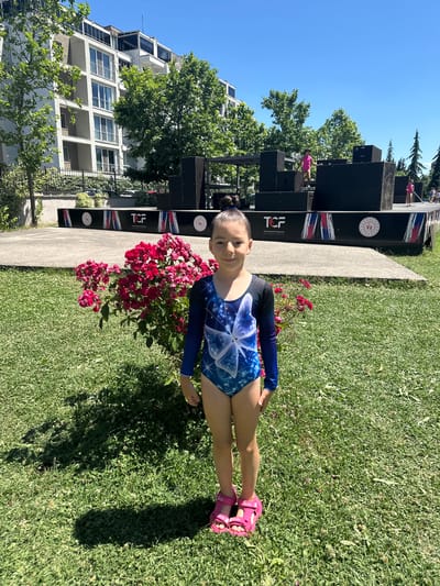 A young girl in a blue and black gymnastics leotard with a butterfly design stands on a grassy area, facing the camera. She is wearing pink sandals. To her left, there is a bed of vibrant red roses. Behind the roses, a concrete path leads to a black structure with various levels and openings. Several other young girls, dressed in pink athletic wear, are visible on and around this structure, suggesting a gymnastics or parkour event. The structure has banners with the text "TCF" and circular emblems. In the background, a modern, white building with multiple floors and windows stands tall, partially obscured by lush green trees. The sky is clear and blue, indicating a bright, sunny day. The scene appears to be outdoors in a park or recreational area, possibly in Sancaktepe, Turkey, given the location context. The girl in the foreground has a slight smile, and the overall atmosphere seems cheerful and active.