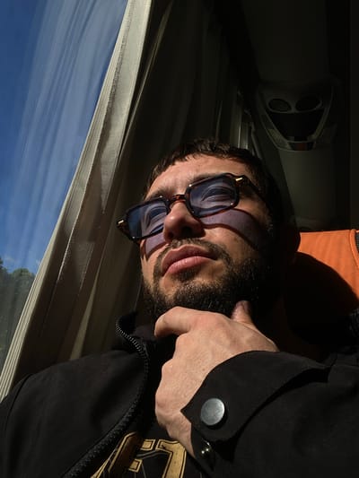 A man is seen from the chest up, wearing sunglasses and a black jacket. He is resting his chin on his right hand. The man is looking to the left, out a window that is covered by a beige curtain. The window shows a bright blue sky with some white clouds and trees. The man's face is illuminated by sunlight, casting a shadow on the right side of his face. The background is dark, with what appears to be part of a bus or train interior visible, including an orange seat. The scene is lit by natural light coming from the window. There is no discernible city or location mentioned in the image or its context.