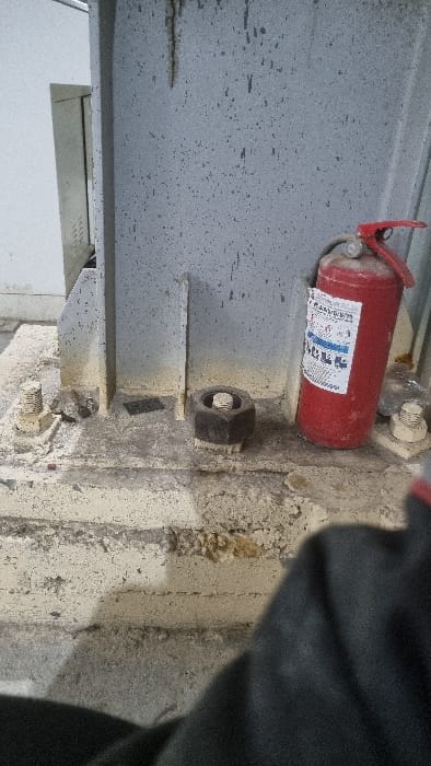 A red fire extinguisher is mounted to a gray, speckled I-beam. A large nut is visible below the extinguisher, secured on a bolt protruding from the beam. More bolts secure the I-beam to the adjacent concrete structure. The area is dusty and appears to be in an industrial or construction environment. The right portion of the image is obscured by a dark, blurry object.