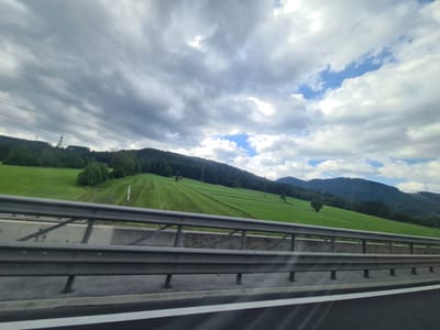 The image is a wide-angle, eye-level outdoor shot of a lush green hillside in Pichlwang, Austria, seen from a highway. The hillside is covered in grass and trees, with evidence of mowed sections indicating agricultural use. A tall electrical pylon is visible on the left side of the hill. The sky above is a mix of dramatic, textured grey clouds and patches of bright blue sky. A metal guardrail runs along the bottom of the frame, suggesting the vantage point is from a vehicle on the highway. There are no people or animals visible in the scene.