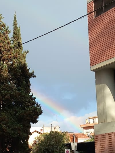 A rainbow is seen in the sky, arching over a street in Drama, Greece. The perspective is from below, looking up between buildings. On the left, a red brick building and a black cable are visible. The right side of the frame shows trees, buildings, and a street sign. The sky is partially cloudy.