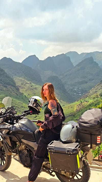 A young woman is sitting on a motorcycle in Lũng Phìn Commune, Vietnam. She is turned toward the camera and has brown hair and a tattoo on her left arm. She is wearing dark pants and a dark tank top, with a dark jacket draped around her shoulders. She is holding a phone in her hands. The motorcycle is black and has luggage attached to the rear. A helmet is sitting on the front of the motorcycle. The background consists of green, mountainous terrain under a partly cloudy sky.
