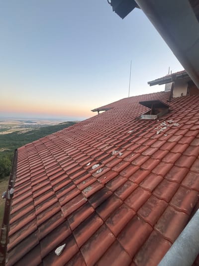 The image depicts a tiled roof made of red clay tiles, angled upwards towards a clear sky. The tiles show signs of age and wear, with some chipped or missing sections and patches of dark discoloration. A metal pipe runs diagonally across the upper right portion of the image, parallel to the roof tiles. Below the roof, a landscape is visible, characterized by a gradient of colors in the sky from light blue to a soft orange and yellow hue near the horizon, suggesting it is either sunrise or sunset. The land below consists of a mix of green trees and fields, with some structures or settlements in the distance. The overall scene appears to be outdoors in a rural or semi-rural setting. There are no people visible in the image. The time of day is likely either dawn or dusk due to the atmospheric lighting. The weather appears clear and calm.