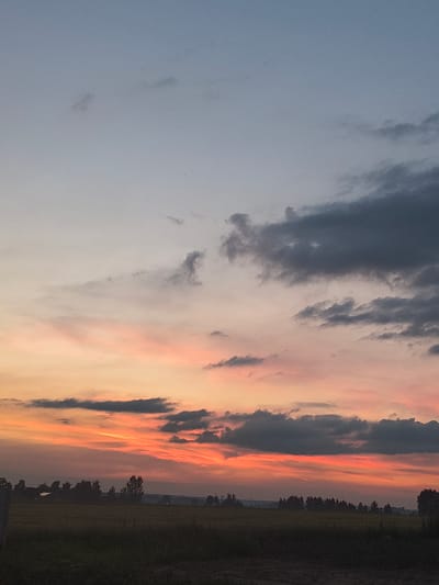 This is a photograph of a sunset over a rural landscape. The sky is filled with clouds, colored in shades of orange, pink, and gray. The sun is not visible, but its light illuminates the clouds, creating a dramatic effect. Below the sky, a dark silhouette of trees and a field can be seen. The location is the city of Galonki, Poland.