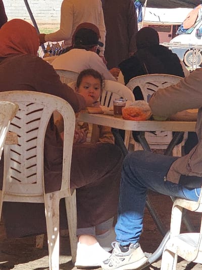 A group of people sits at a table with white chairs. A baby lies in a chair, looking at the camera. People surround the table, and there are drinks and food on the table. One person wears jeans and sneakers. Another person wears a dark-colored shirt. Several people wear brown clothing. A van is visible in the background.