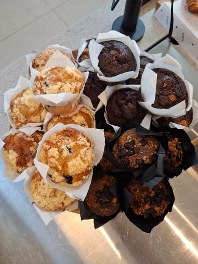 The image displays an overhead view of a diverse selection of muffins arranged on a metallic, reflective display counter within a commercial establishment in Bnei Dror, Israel. Approximately 20-25 muffins are visible, grouped by type. To the left and top-left, several light-colored muffins with a crumble topping, some containing visible blueberries, are presented in white pleated paper liners. The central and upper-right areas feature multiple dark chocolate muffins, each topped with chocolate chips, also housed in white pleated paper liners. The lower-right portion shows darker-hued muffins, possibly coffee or nut-flavored, adorned with chocolate chips and granular pieces, contained within black pleated paper liners. The metallic counter surface exhibits reflections of overhead lighting. In the upper-right background, a portion of a white cardboard box with blue text and a partially visible croissant are discernible. A black vertical fixture is present in the upper-middle background. No individuals or specific actions are depicted.