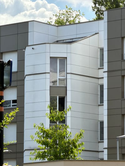 The image captures a segment of a modern building's facade, characterized by its angular design and metallic cladding. The building features several windows, some with visible reflections of trees and sky. Lush green foliage from nearby trees partially obscures the building, suggesting an outdoor setting with abundant vegetation. The sky appears bright with large, fluffy clouds, indicating daytime, likely sunny weather.
The composition is vertical, with the building occupying the majority of the frame. The architectural style is contemporary, with clean lines and a mix of light-colored panels that create a geometric pattern. One prominent window shows a clear reflection of the sky and clouds, while others reflect the surrounding greenery.
The overall impression is one of urban architecture set against a natural backdrop. The sunlight filtering through the leaves and reflecting off the building's surfaces creates a dynamic play of light and shadow. No people are visible in the image. The scene appears peaceful and serene, with no specific activity taking place. The location context is identified as Évry-Courcouronnes, France. There is no visible text in the image.