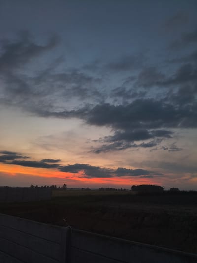 This image captures a dramatic sunset over a rural landscape, likely on the outskirts of Galonki, Poland. The sky is a spectacle of color, transitioning from deep, bruised blues and grays at the top to vibrant oranges and reds near the horizon. Dark, heavy clouds are scattered across the upper sky, while thinner, wispy clouds are illuminated by the setting sun lower down.

The foreground is dominated by a dark, silhouetted field and what appears to be construction elements. A substantial concrete fence runs horizontally across the lower part of the frame, appearing weathered and textured. Beyond the fence, the ground is rough and uncultivated, with areas of dark soil and shadow.

In the mid-ground, a line of trees creates a dark, undulating silhouette against the vibrant sky. These trees vary in density, with some appearing as dense stands and others as more sparse arrangements. The horizon line is low, emphasizing the expansive sky. The overall impression is one of a serene, yet powerful natural event unfolding in a developing or undeveloped rural setting.